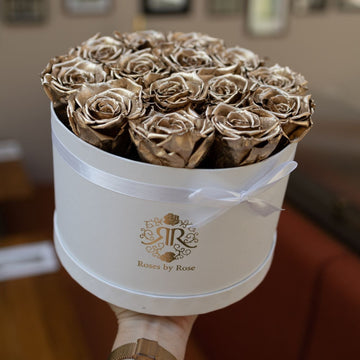 White round box with gold roses and 'Roses by Rose' branding against a blurred indoor background.